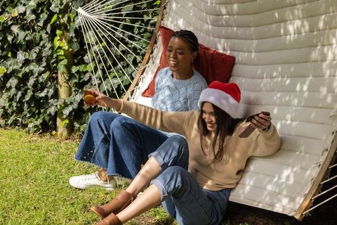 Diverse friends lounging on hammock in backyard wearing santa hat and using smartphone