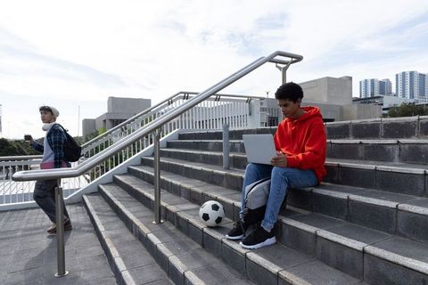 Multicultural students studying on campus steps using laptop and phone with soccer ball