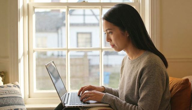 Focused young woman working remotely on laptop by sunlit window in cozy minimal home