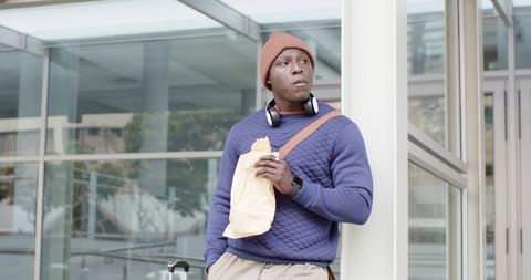 African American commuter leaning on column eating takeaway with suitcase and headphones