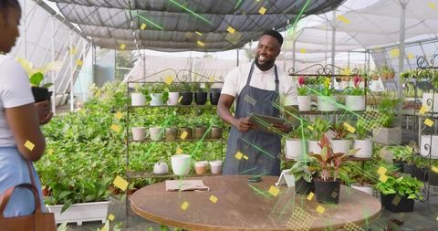 Smiling gardener vendor helping customer holding tablet among potted plants in greenhouse
