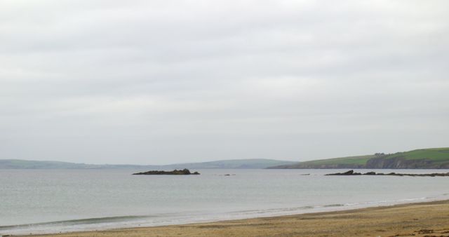 Image depicts a quiet beach with a calm seascape on a cloudy day, showing sandy shore leading to tranquil ocean under an overcast sky. Ideal for promoting travel to peaceful coastal areas, usage in self-care and relaxation content, showcasing nature's beauty, or featuring in blogs about solitary retreats and meditative environments.