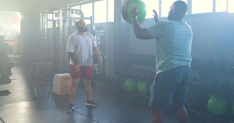 African men tossing green medicine ball during partner workout in backlit industrial gym
