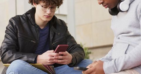 College friends checking phones on campus steps with notebooks and headphones, studying