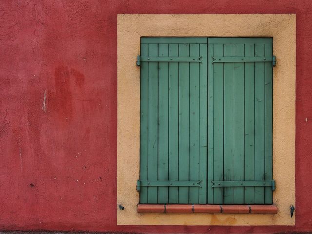 Green wood window shutters are set against a red wall with a cream border. It evokes a rustic and vintage feel, typical of Mediterranean architecture. Suitable for architectural studies, design inspiration, or as a decorative element for home and garden publications.