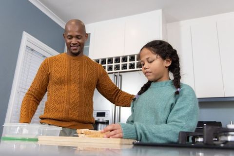 Father guiding daughter preparing sandwich on kitchen island in cozy sweaters