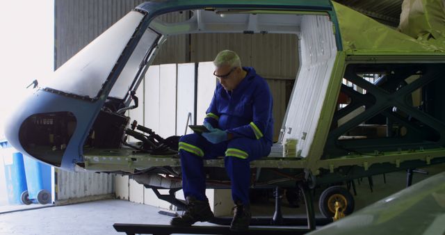 Engineer in blue overalls and safety glasses is using a tablet while seated inside a helicopter fuselage in a maintenance hangar. This setting is indicative of aerospace and aviation industry activities such as inspection, repair, and maintenance operations. Useful for illustrating themes of engineering, technology usage in aircraft maintenance, and industrial safety protocols.