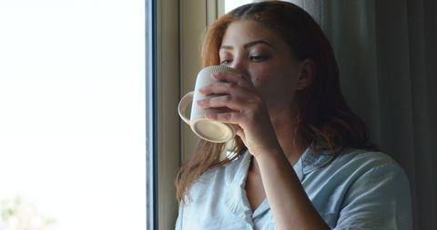 Contemplative woman wearing blue shirt sipping coffee by window holding ribbed mug