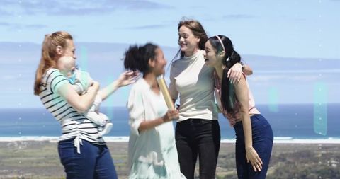 Group of women laughing on ocean overlook holding baby and rolled scroll, celebrating outdoors