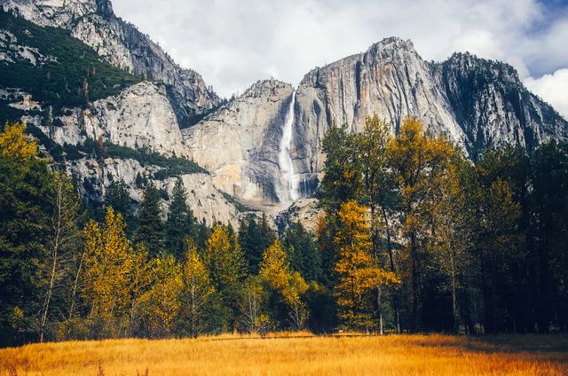 This breathtaking scene captures a majestic waterfall cascading down rugged cliffs amidst an autumn forest in Yosemite National Park. Golden leaves on the trees contrast beautifully with the gray rock faces and adds to the picturesque natural beauty. Ideal for promoting travel destinations, nature conservation, adventure activities, and stock images related to the great outdoors.