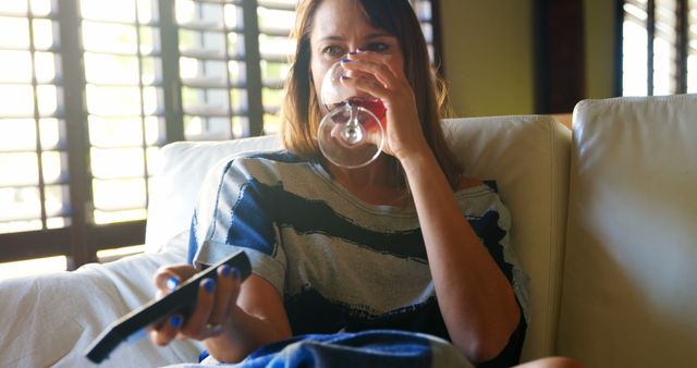 Woman sitting on couch enjoying a glass of wine while watching TV holding remote control. Ideal for depicting relaxation at home, leisure activities, or independent lifestyle content. Can be used in lifestyle blogs, advertisements for home goods, wine promotions, or articles about solo hobbies.