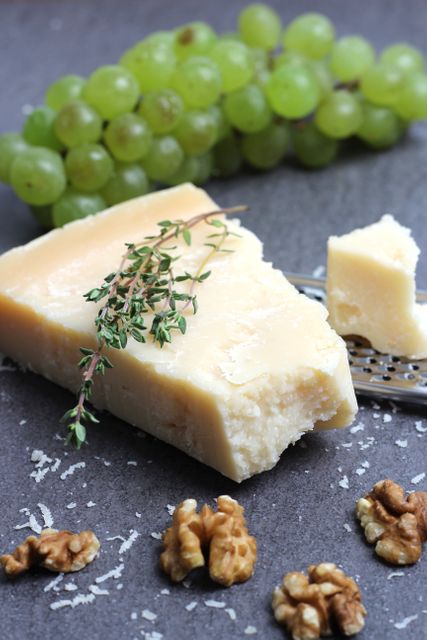 A piece of parmesan cheese with thyme, a bunch of green grapes, and scattered walnuts on a dark textured surface. Ideal for illustrating gourmet food, cheese pairing, Italian cuisine, or wine tasting events. Use in blogs, presentations, or menus for restaurants or culinary websites.