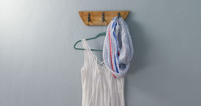 White summer dress and lightweight scarf hanging on rustic wooden hook against blue wall. Ideal for illustrating fashion trends, home organization tips, minimalist decor ideas or wardrobe essentials in lifestyle blogs and magazines.