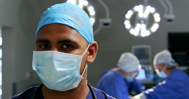 Focused young surgeon wearing surgical mask and cap in an operating room with other surgeons performing surgery in the background. Useful for healthcare narratives, medical training materials, healthcare advertisements, and health-related articles.