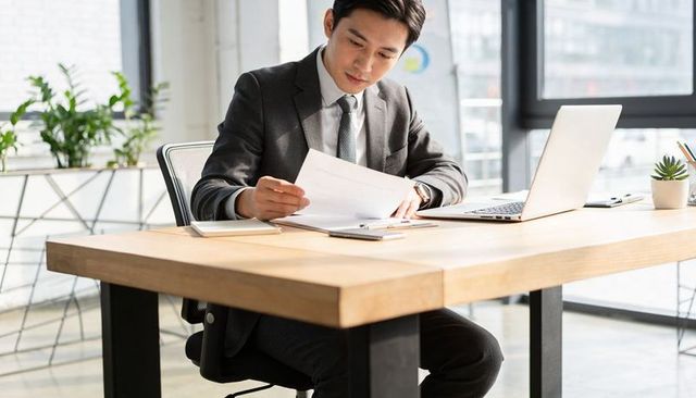 Reviewing documents at modern wooden desk with open laptop in bright urban office