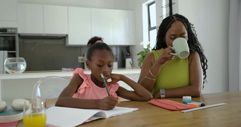 African american mother supervising daughter doing homework at kitchen table while drinking coffee