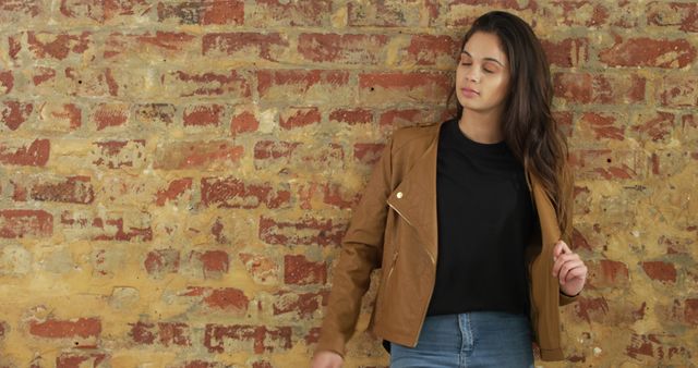 Young woman standing confidently against a rustic brick wall while wearing a brown jacket and jeans. Perfect for fashion websites, articles about confidence, lifestyle blogs, or urban-themed advertisements.