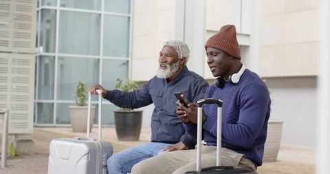 African American Senior and Young Man Waiting with Luggage Using Smartphone Outdoors