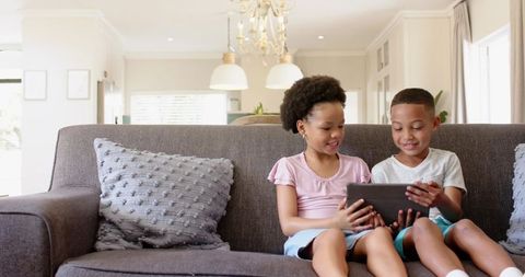 Diverse Siblings Sharing Tablet on Cozy Sofa in Modern Living Room