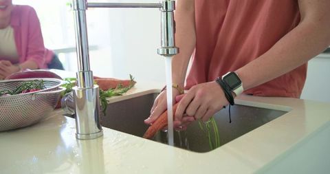 Washing fresh carrots under chrome faucet in modern kitchen preparing healthy meal