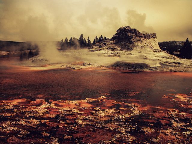 Steaming geyser landscape surrounded by dramatic sky. Dark, earthy tones with rich natural textures dominate the scene. Perfect for use in travel brochures, educational material on geothermal activity, or posters capturing the beauty of nature and national parks.