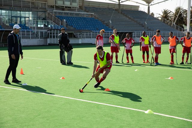 Multi-ethnic group of teenage male field hockey players, practiicing before a game, standing with their coach in a pitch, one player hitting ball with hockey stick. Sport game competition.