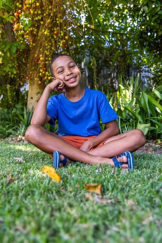 African american boy sitting cross-legged on lawn under fruit tree, smiling outdoors