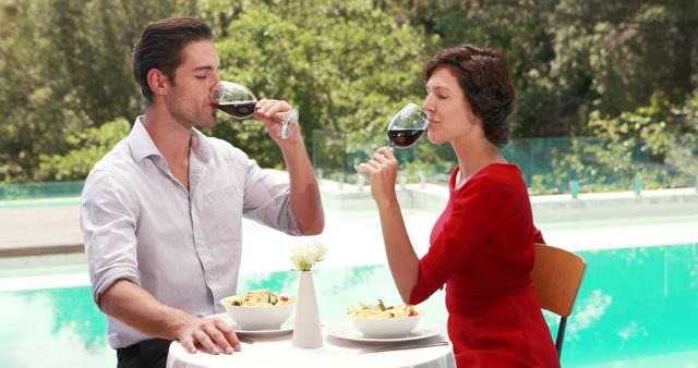 A Caucasian couple enjoys a romantic meal outdoors by a pool, with copy space. They are toasting with glasses of wine, creating a relaxed and intimate dining experience.