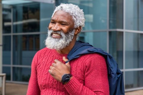 Mature african american man smiling in red sweater carrying navy jacket near glass building