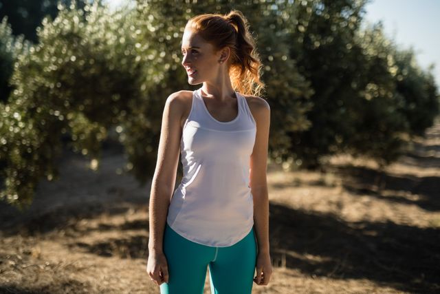 Young woman in athletic wear standing in a sunlit field, smiling and looking away. Ideal for use in fitness, health, and lifestyle promotions, as well as outdoor and nature-related content.