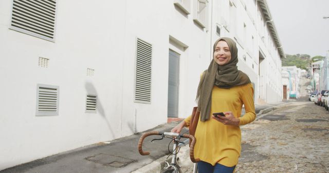 Happy biracial woman in hijab walking in city street wheeling bike and using smartphone. City living, communication, transport and modern urban lifestyle.