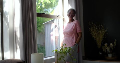 Senior woman smiling by sunlit window with indoor plant and soft curtain reflection