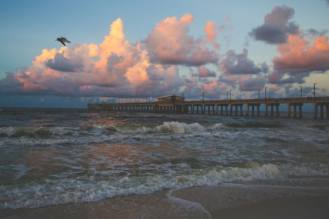 This image shows a beautiful sunset over a coastal pier with waves crashing on the shore and colorful clouds in the sky. Ideal for travel blogs, nature websites, or beach resort promotions, conveying a sense of peace and natural beauty.