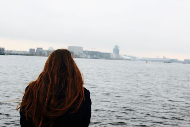 A woman with long hair is staring at a calm river on a cloudy day, providing a sense of solitude and peaceful contemplation. The urban cityscape is visible in the distance, adding to the serene atmosphere. This could be used to represent themes of introspection, solitude, and urban living. Ideal for articles or advertisements focusing on mental health, urban exploration, or travel.