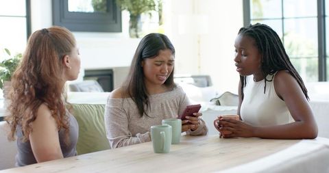Diverse female friends comforting distressed woman over smartphone in sunny living room