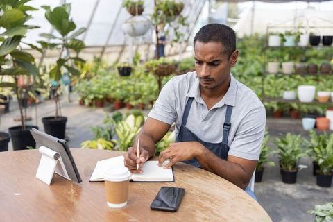 Greenhouse nursery worker writing inventory notes using tablet and planning plant orders