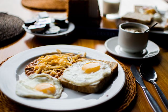 Close-up of delicious breakfast featuring fried eggs, toast, baked beans topped with shredded cheese, and a cup of coffee. Ideal for food blogs, restaurant menus, or nutrition and lifestyle articles emphasizing hearty morning meals.