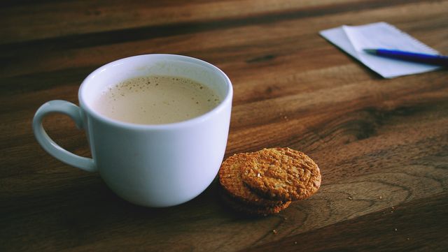 Cup of steaming coffee placed beside two oatmeal cookies on rustic wooden table. Background includes notepad with pen. Perfect for illustrating cozy moments, morning routines, or work breaks in cafes and home offices.