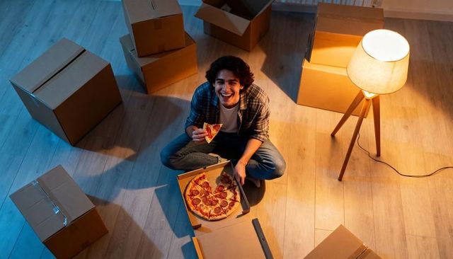 Young man eating pizza while unpacking boxes during move into apartment at night