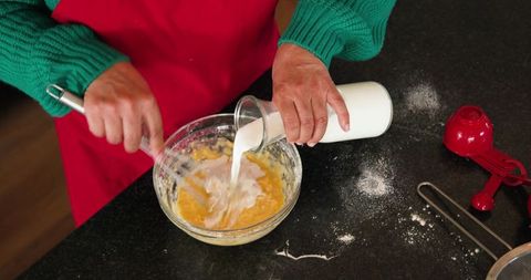 Mature woman pouring milk and whisking batter in glass bowl on dark countertop