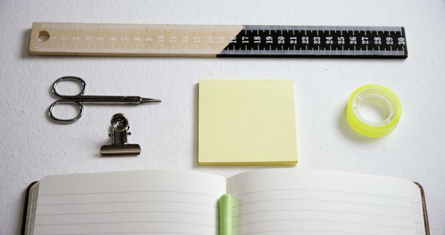 An assortment of office supplies including a ruler, scissors, sticky notes, a binder clip, and an open notebook is neatly arranged on a white surface, with copy space. These items are typically used for organization and productivity in a work or academic setting.