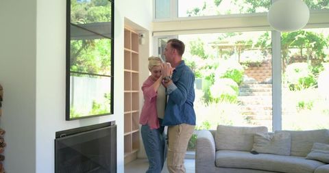 Romantic couple dancing in sunlit modern living room by floor-to-ceiling windows