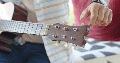 Two mature men tuning acoustic guitar on couch, close-up hands and headstock