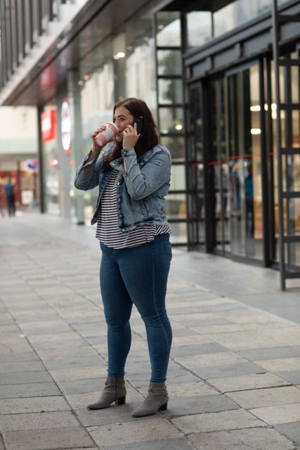 Curvy Caucasian woman enjoying a takeaway coffee while talking on her smartphone in an urban setting. She is dressed in a casual outfit with a denim jacket and jeans, standing in front of a modern building. This image is ideal for use in articles or advertisements related to urban lifestyle, multitasking, modern living, or fashion for curvy women.