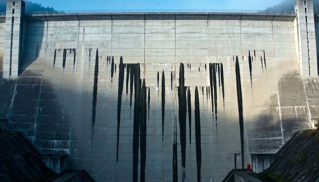 Displaying concrete dam wall with vertical seepage stains, crest railing and red canister
