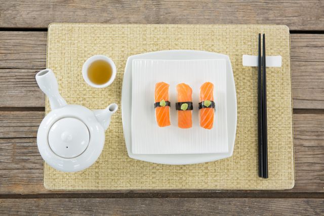 Top view of traditional Japanese sushi with salmon, wasabi, and soy sauce on a white plate, accompanied by a teapot and cup of tea. Black chopsticks are placed on a woven mat against a rustic wooden table. Ideal for use in food blogs, restaurant menus, culinary magazines, and advertisements promoting Japanese cuisine.