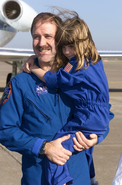 Astronaut in a blue NASA flightsuit holding child at a crew return ceremony at an airport. Ideal for use in contexts related to space exploration, parent-child bonding, aviation events, and governmental programs. Could be used in articles about astronaut life, promotional material for family-oriented aerospace events, or educational content for children about space missions.