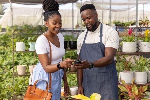 African american customer using smartphone with plant nursery owner in greenhouse