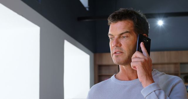 Man standing in a modern office, talking on a smartphone. He appears focused on the conversation. Ideal for use in business communication, technology advertisements, or illustrating a professional environment.