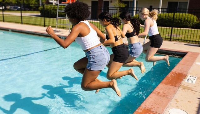 Four friends jumping into pool in summer wearing swimwear and casual denim shorts